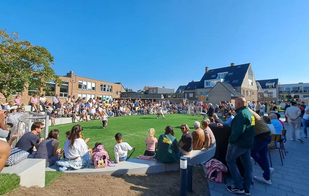 Ouders, kinderen en buurtbewoners volgen een activiteit op het plein met kunstgrasveld en zittribunes